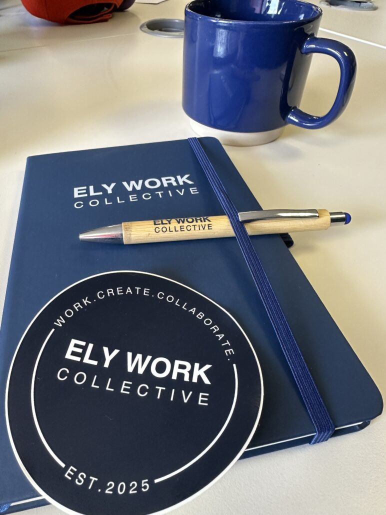 Ely Work Collective branded notebook, pen and sticker reading "Work. Create. Collaborate. Est. 2025", with a blue mug on a desk