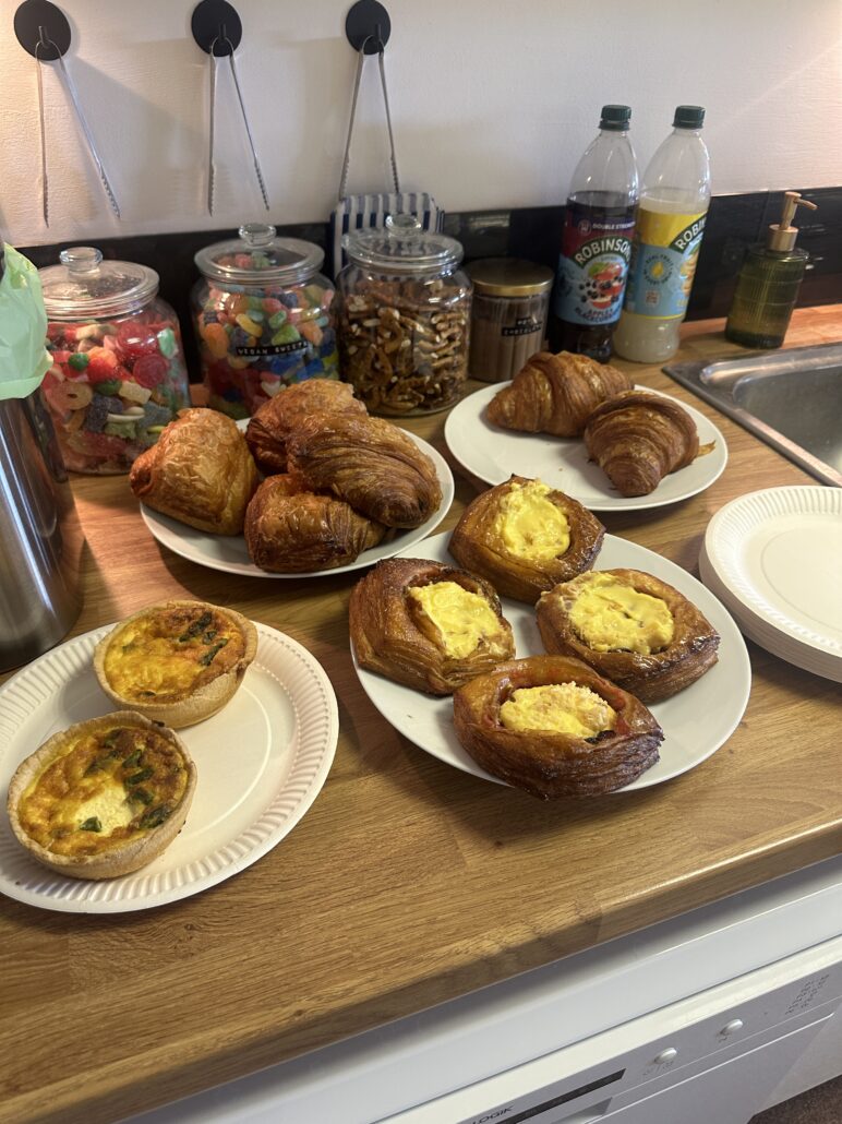 Plates of croissants, custard pastries and mini quiches on a kitchen counter, alongside jars of sweets and nuts at Ely Work Collective